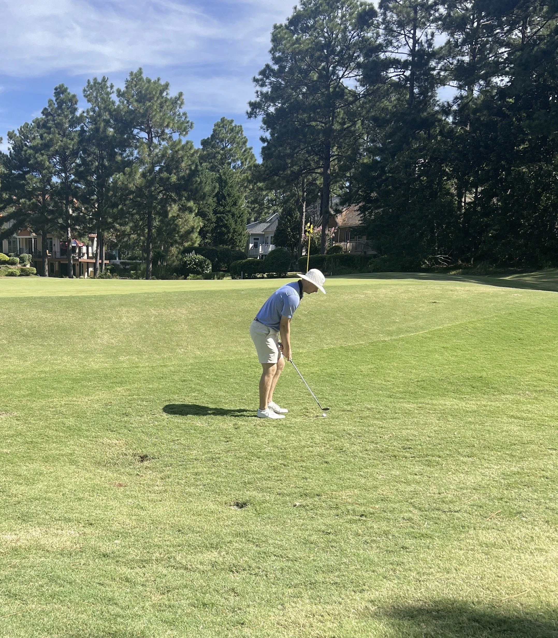 A man in a white hat, blue shirt, and beige shorts playing golf on a grassy course.