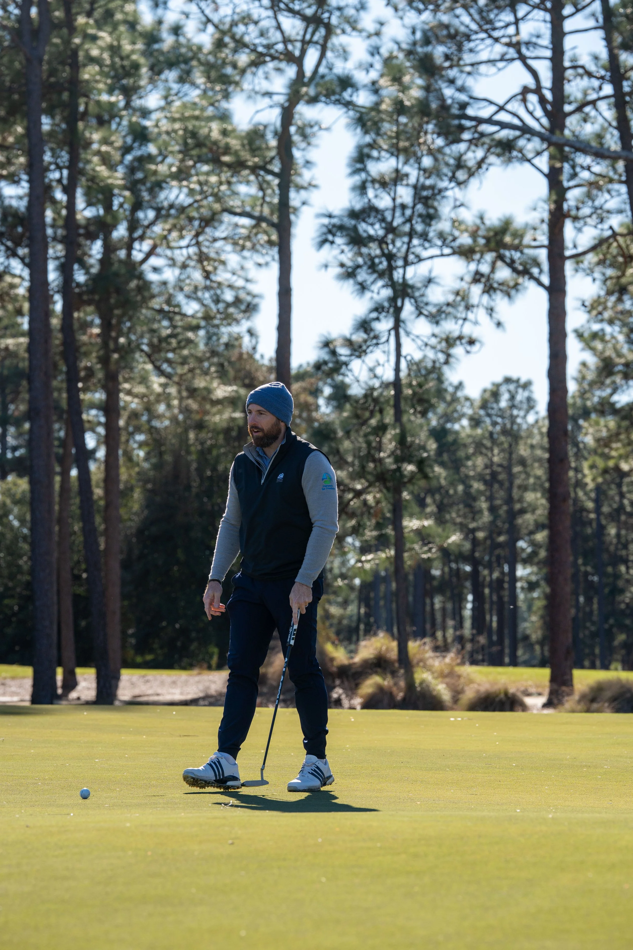A man playing golf on a green, holding a golf club, with trees and clear sky in the background.