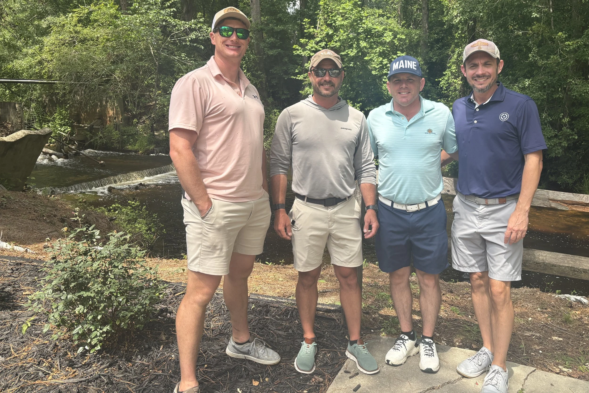 Four men standing outdoors near a small river in a wooded area, dressed in casual golf attire, smiling at the camera.