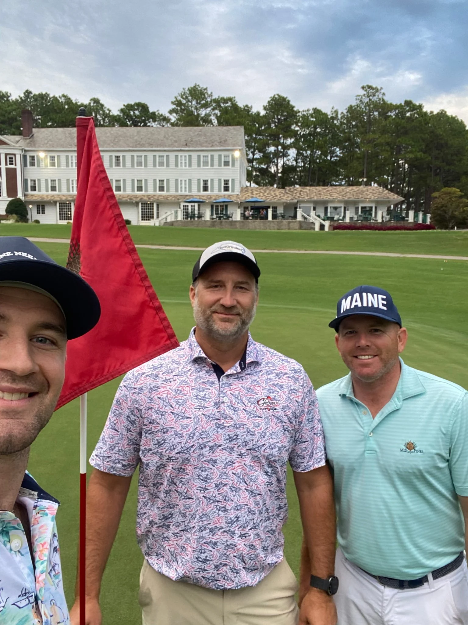Three men standing on a golf course, holding a red flag, with a large white house and trees in the background, wearing casual golf attire including hats.