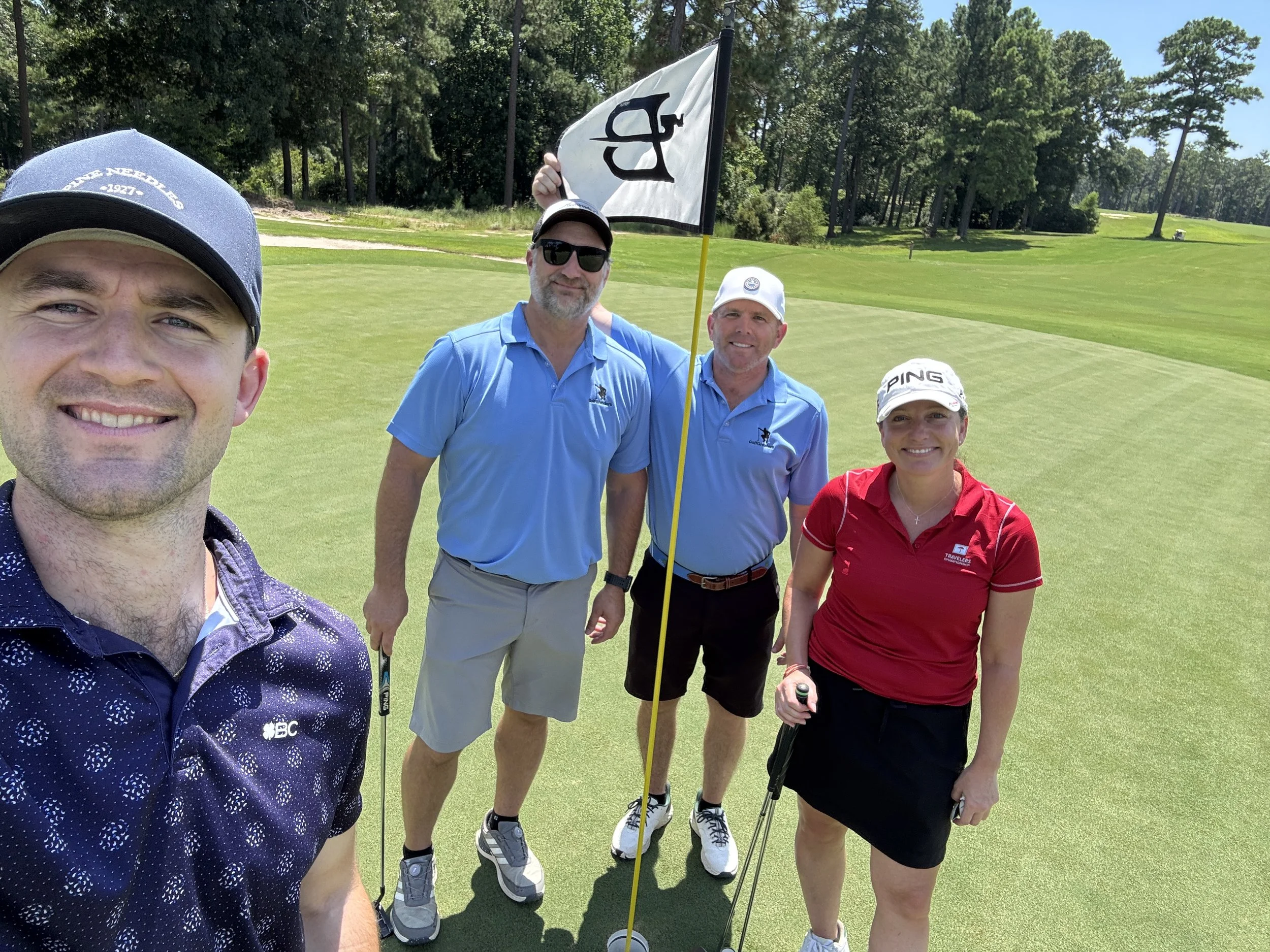 Four people on a golf course with trees in the background, taking a selfie near the green, with one person holding a golf flag and all holding golf clubs.