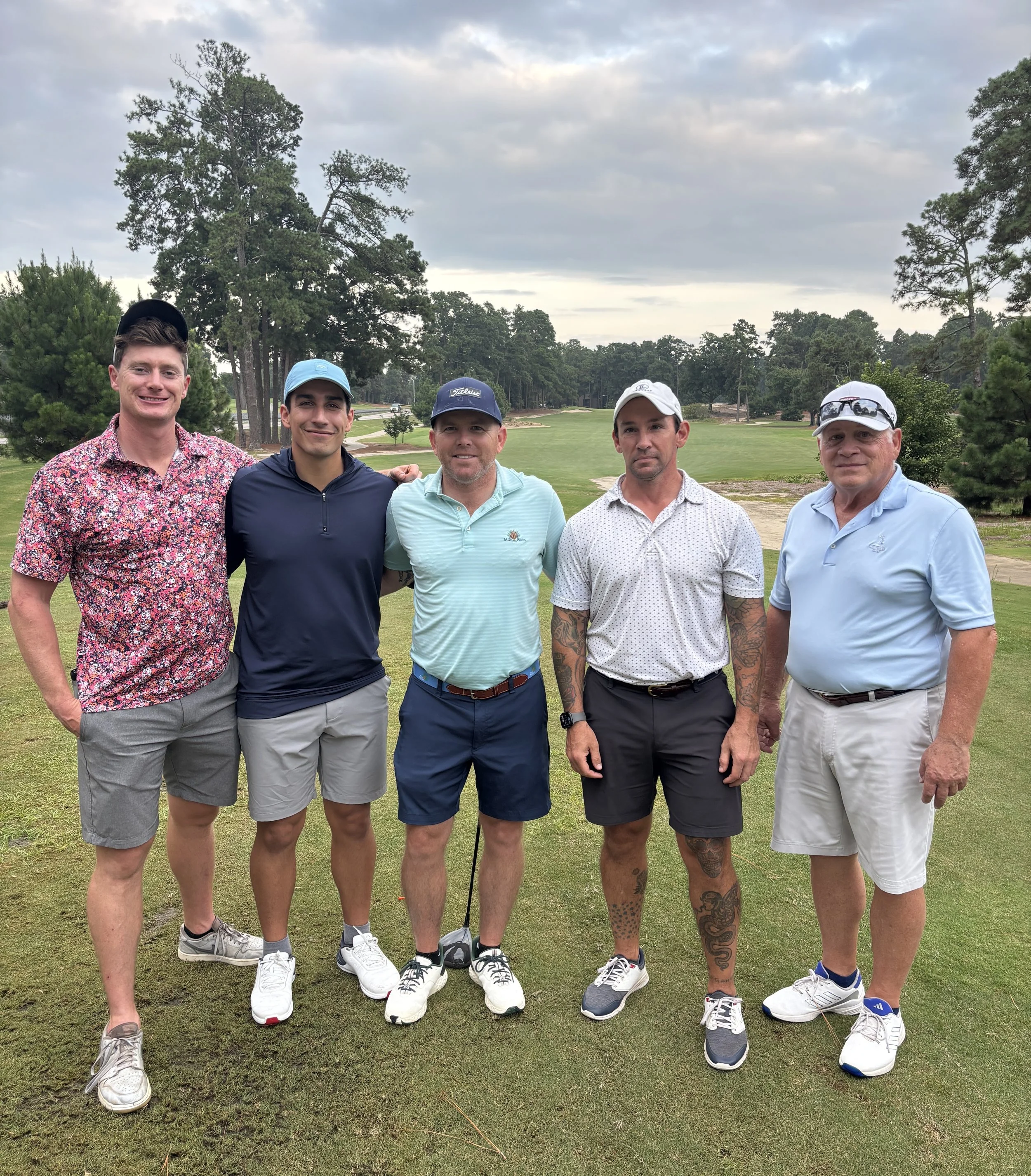 Group of five men standing together on a golf course, dressed in golf attire, with trees and a cloudy sky in the background.