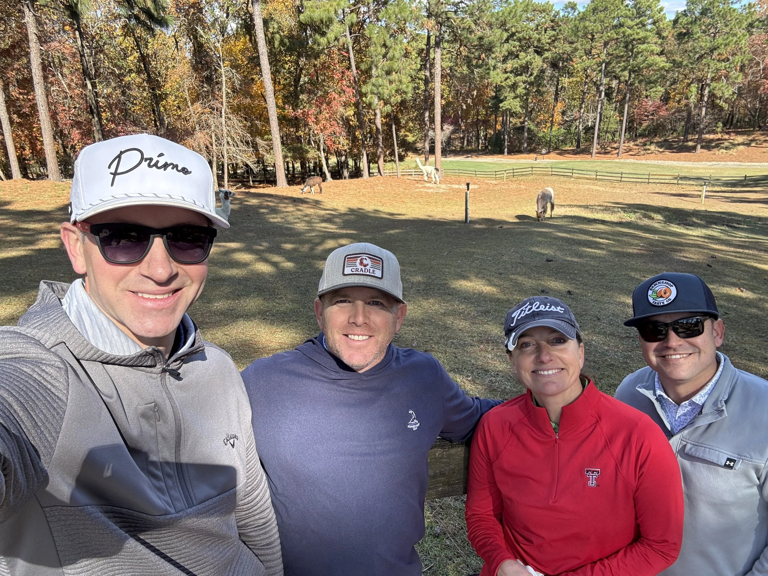 Four people taking a group selfie outdoors on a golf course with fall-colored trees and grazing horses in the background.