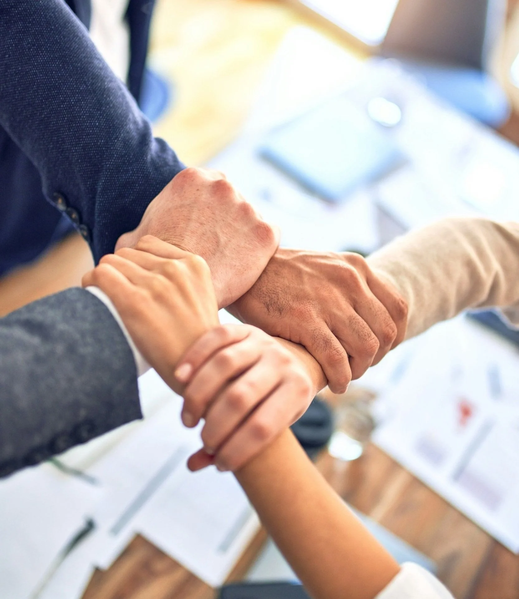 Multiple people holding hands in a team-building gesture, seen from above, with a meeting table and documents in the background.