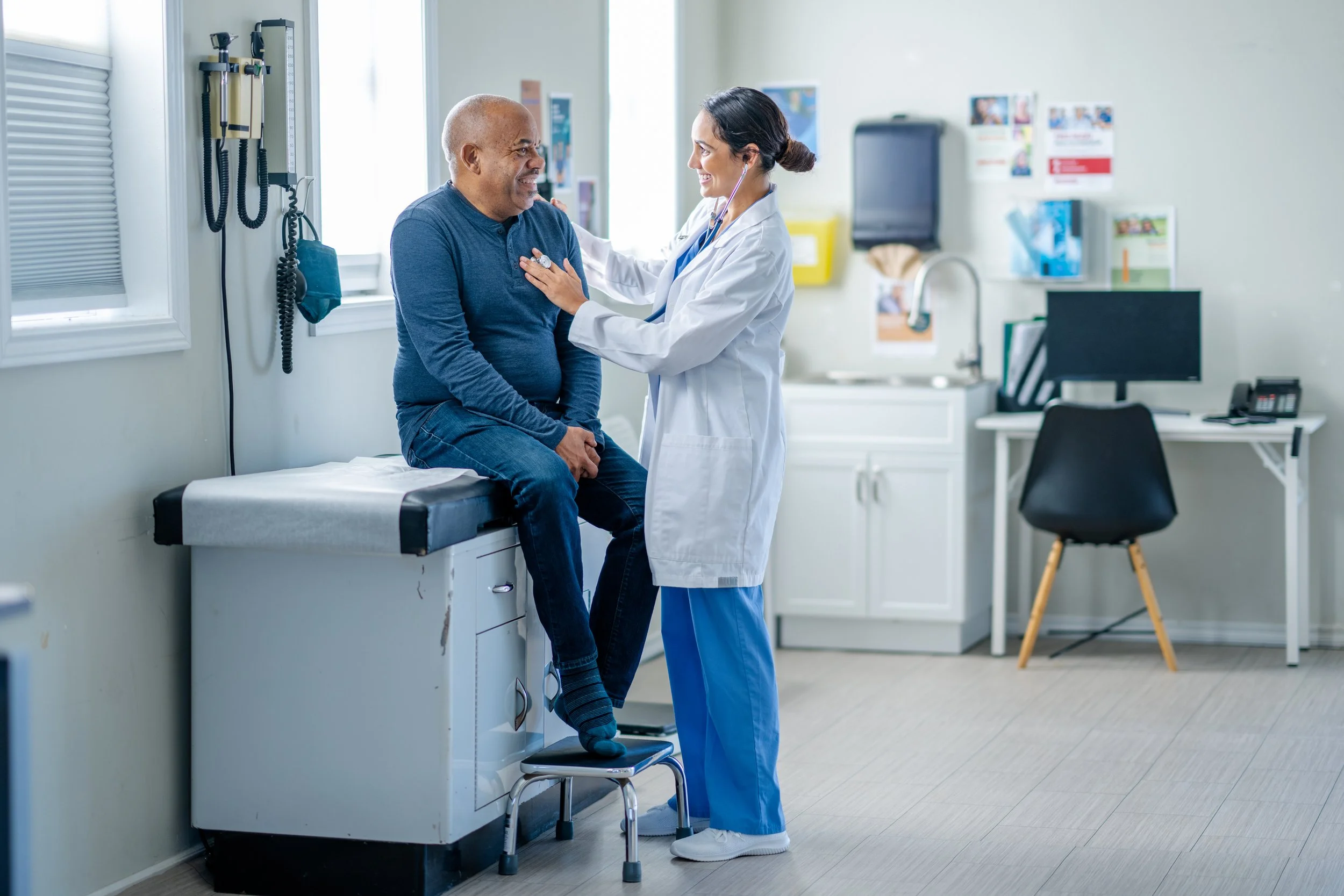 A physician wearing blue scrubs and a white lab coat cares for a patient wearing blue jeans and a blue long-sleeve T-shirt