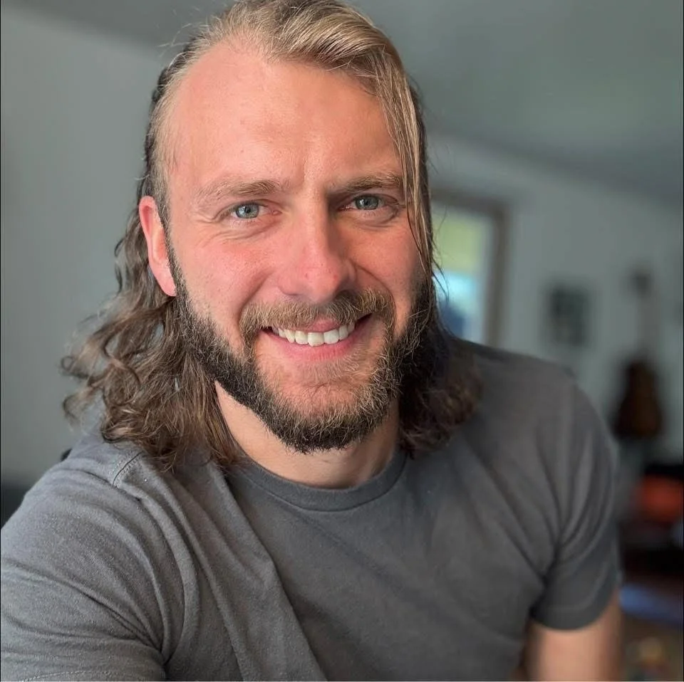 A man with long brown hair, a beard, and blue eyes smiling at the camera, wearing a gray t-shirt, inside a home with a blurred background of a window and furniture.