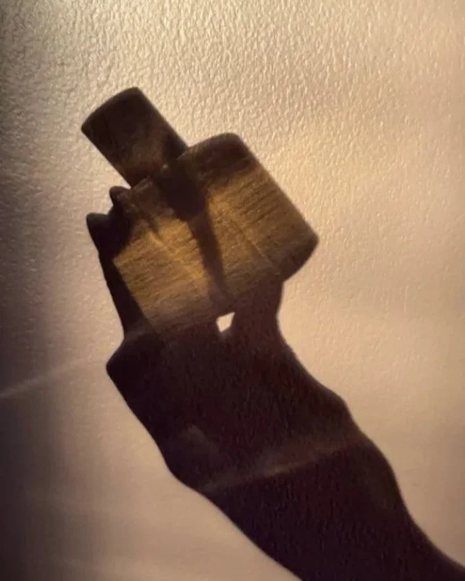 A close-up of a dark-colored hand holding a small wooden object against a plain beige wall, with dramatic lighting creating shadows.