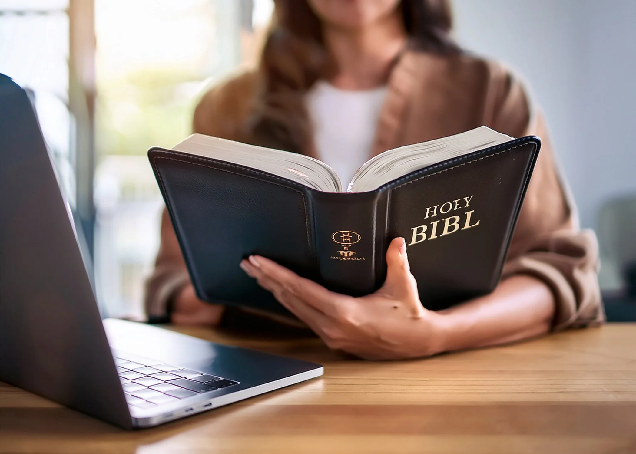 A woman sitting at a wooden table holding an open black Bible with the words 'HOLY BIBLE' on the cover. There is a silver laptop on the table next to her, and she is wearing a brown jacket and white shirt. The background is blurred with natural light coming through a window.