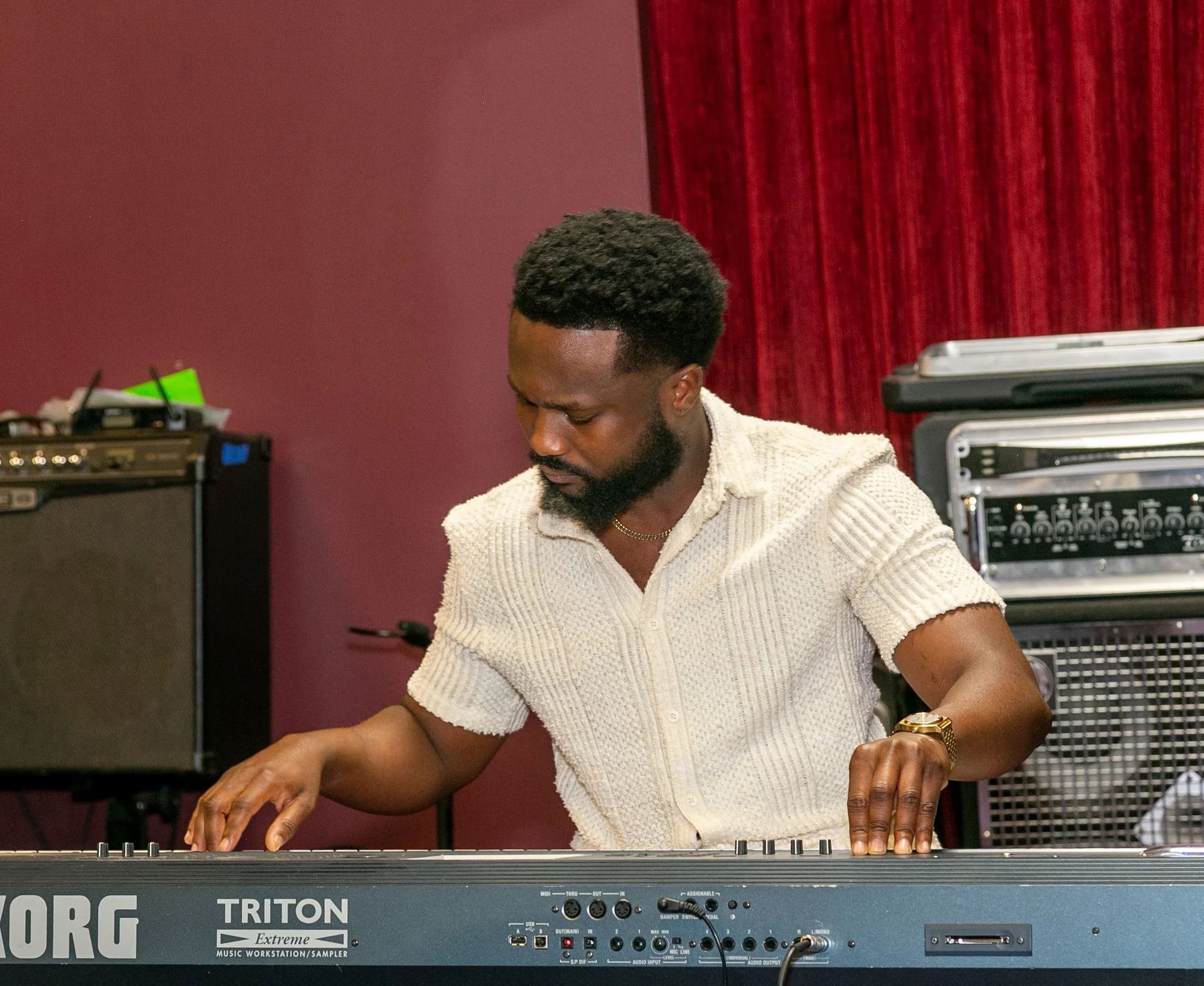 A man playing a keyboard in a room with musical equipment and red curtains.