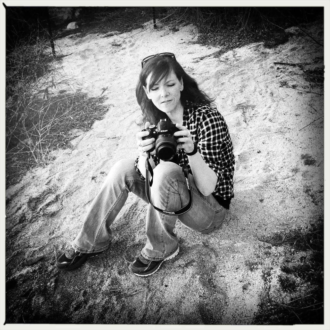 A woman crouching on a sandy trail, holding a Nikon camera and looking at it, surrounded by sparse vegetation.