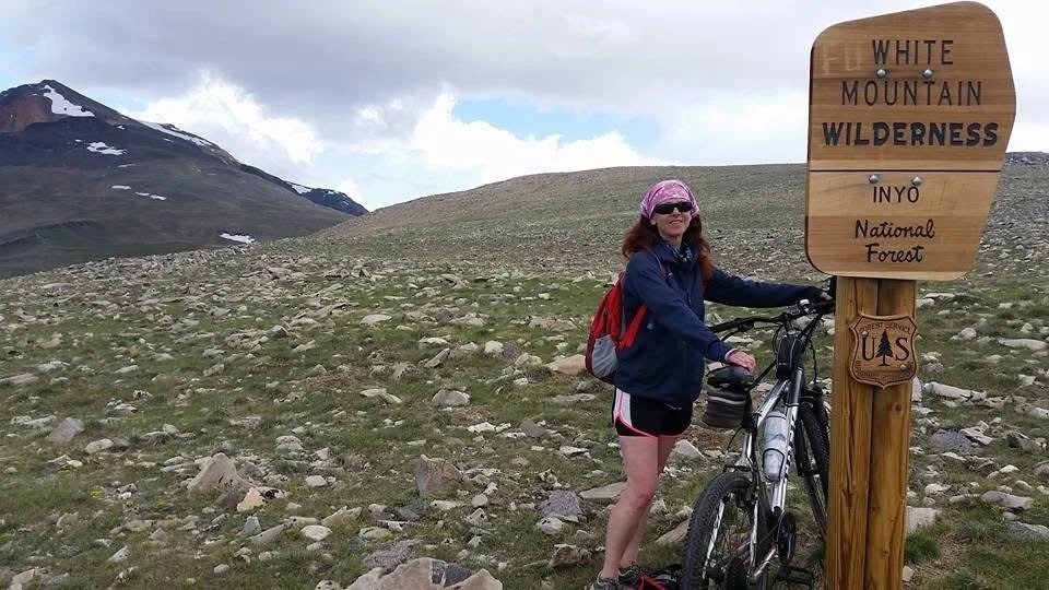 A woman with a pink bandana, sunglasses, a blue jacket, and shorts standing next to a bicycle at White Mountain Wilderness, Inyo National Forest, with a scenic mountain landscape in the background.