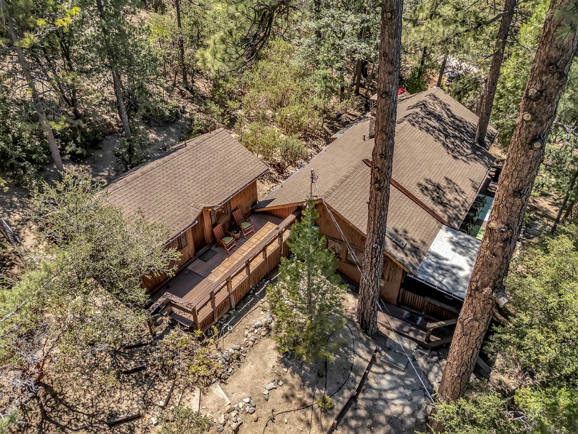 Aerial view of a wooden house and a smaller shed surrounded by tall pine trees in a forest, with a deck and outdoor chairs visible.