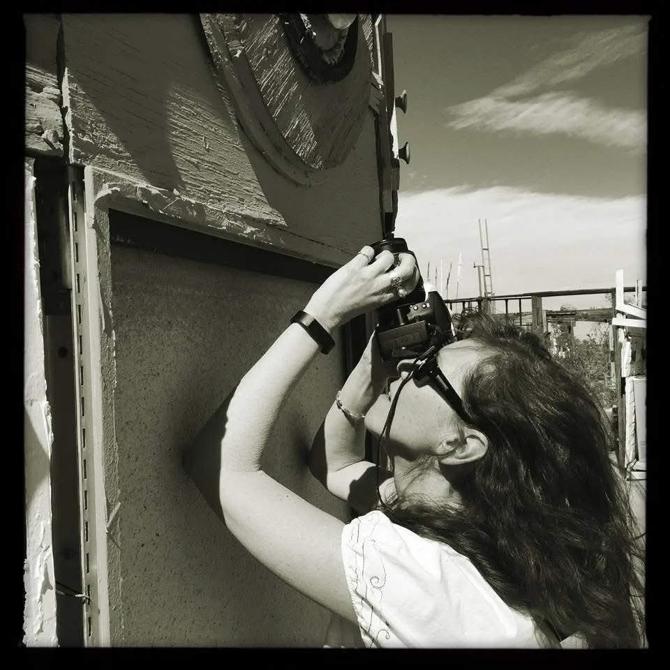 A woman with dark wavy hair and glasses is taking a photo of a building with a camera. She is standing outdoors on a balcony or rooftop, with her eye close to the camera's viewfinder, and appears focused on capturing the shot.
