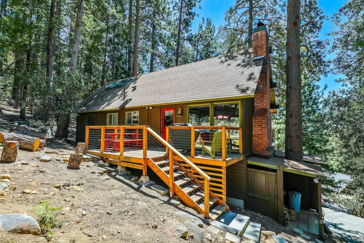 A small house with a beige roof and red brick chimney sits among tall pine trees. It has a wooden deck with stairs leading up to a red door and outdoor furniture.