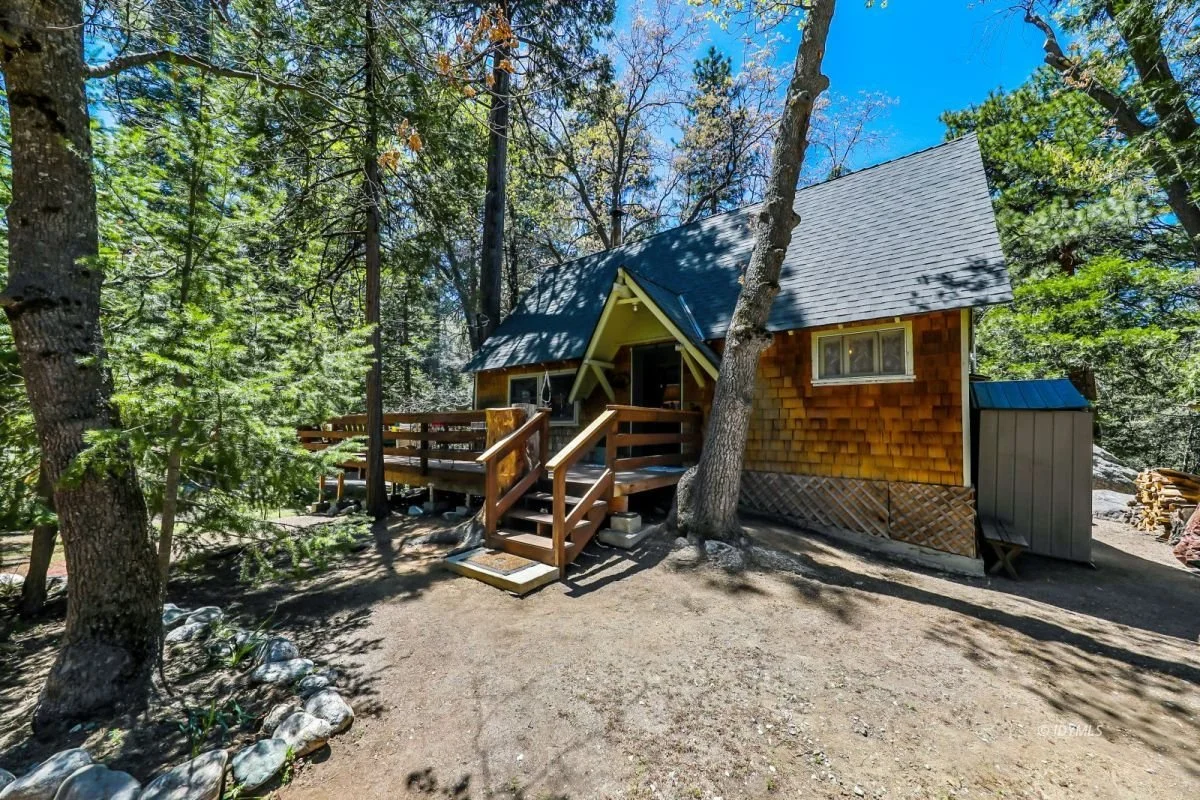 A small wooden cabin with a sloped roof, located in a wooded area with tall trees and a dirt ground. The cabin has a front porch with stairs and a railing.