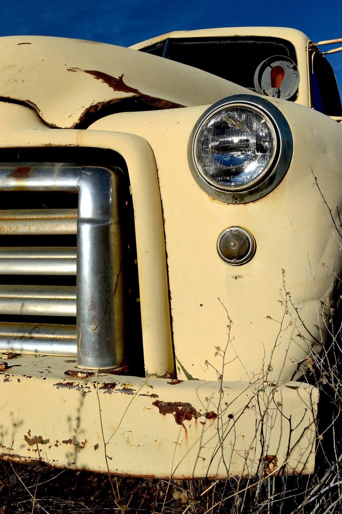 Close-up of an old, yellow, rusty truck with damaged hood, headlight, and grille, surrounded by dry grass and weeds under a clear blue sky.