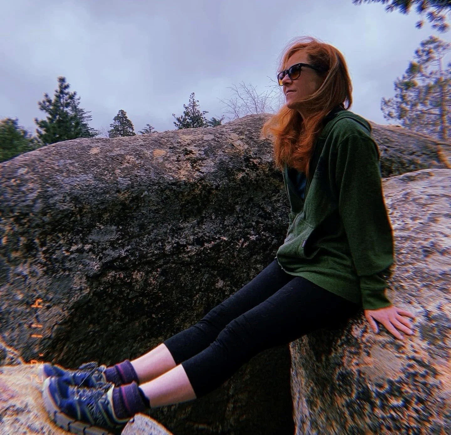 A woman with red hair and sunglasses sitting on a large rock outdoors, wearing a green jacket, black pants, and hiking shoes.