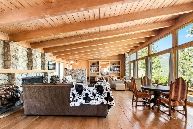 Living room with wooden ceiling, stone fireplace, large windows, and mixed furniture including a sofa with a cowhide throw, armchairs, and a round dining table with chairs.
