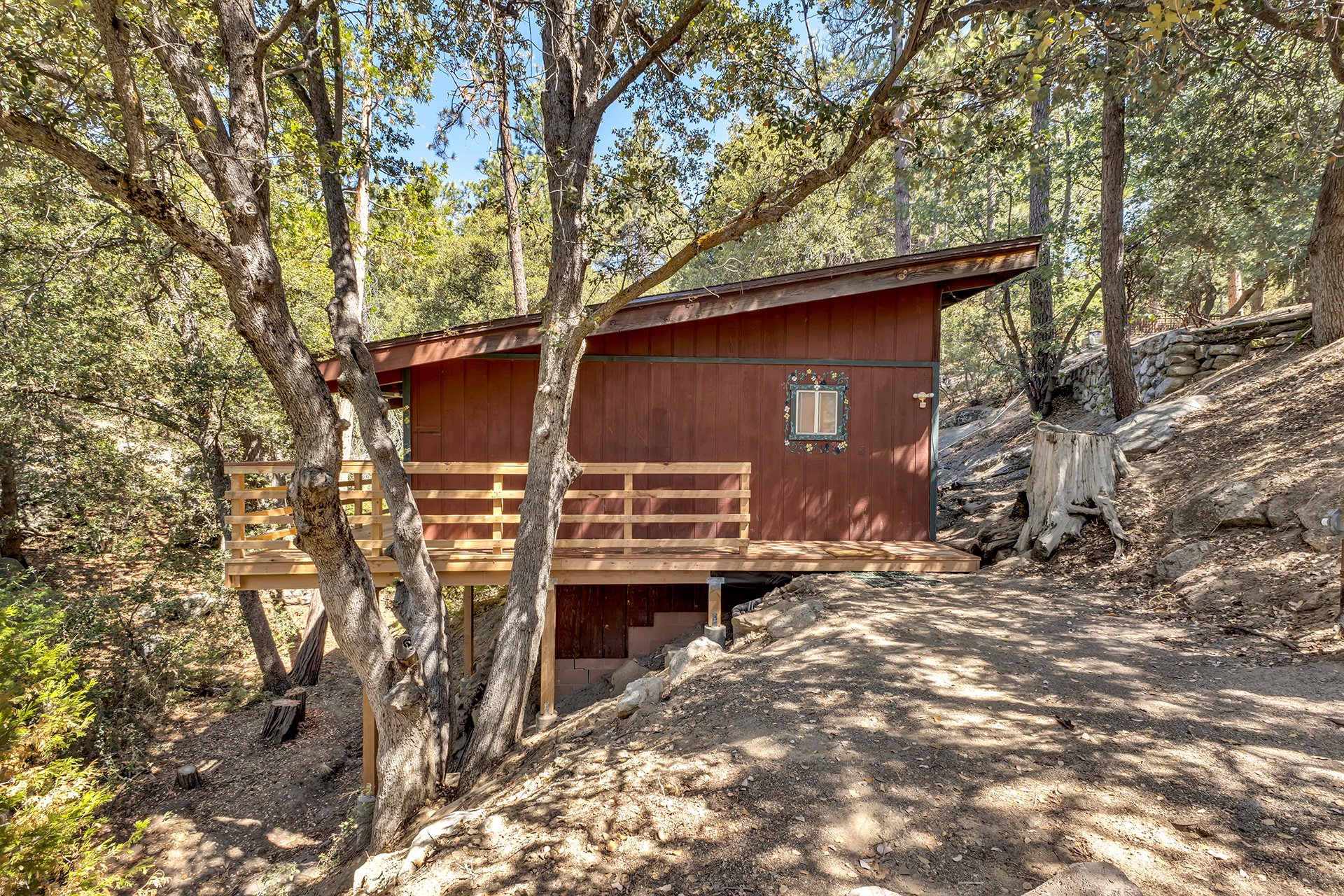 A small house with a sloped roof and a wooden deck built on a hillside surrounded by trees and rocky terrain.