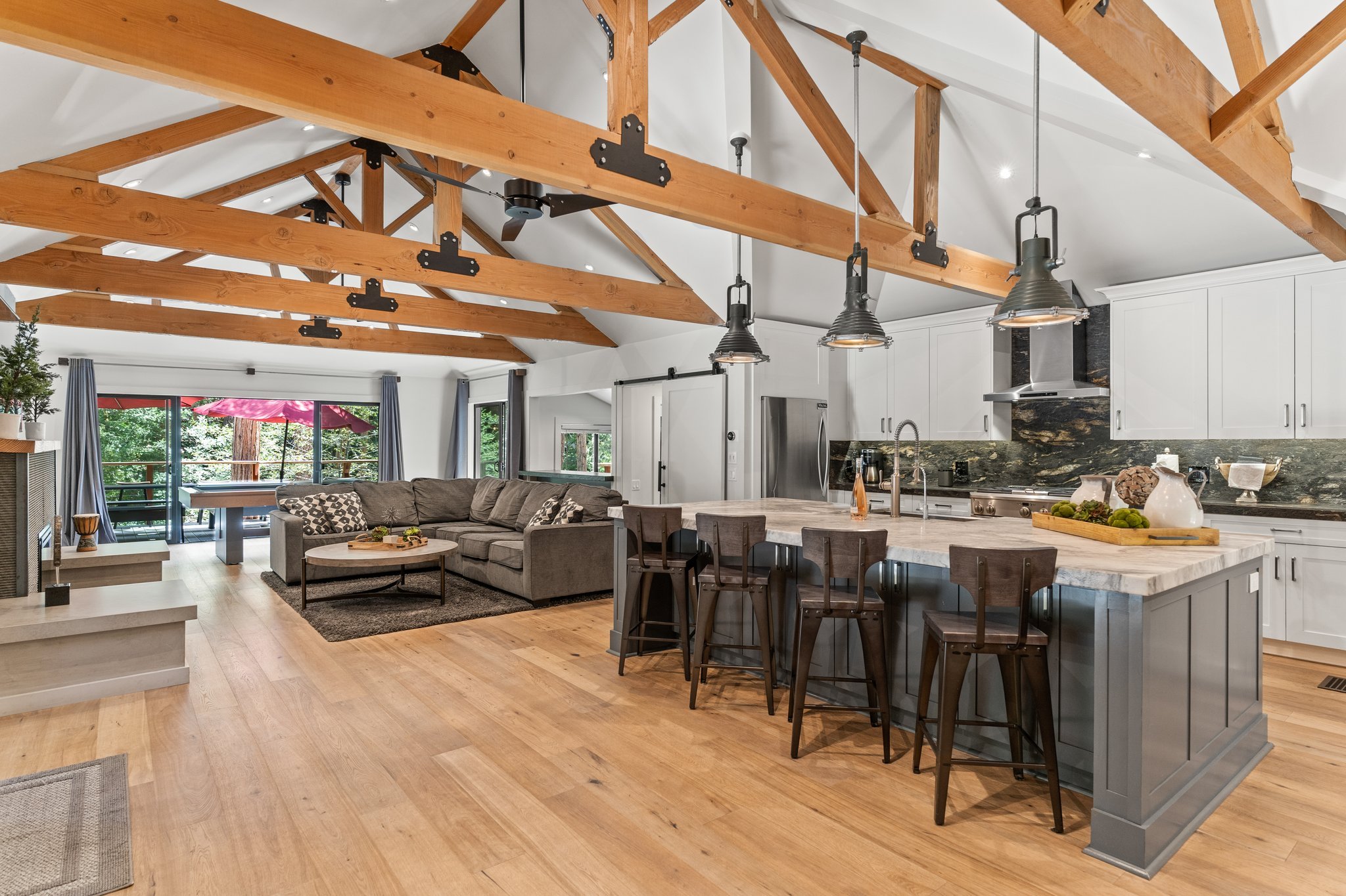 Open-concept living room and kitchen area with exposed wooden beams on the ceiling, a large gray sectional sofa with patterned pillows, a kitchen island with chairs, stainless steel refrigerator, and sliding glass door leading to a balcony with trees outside.