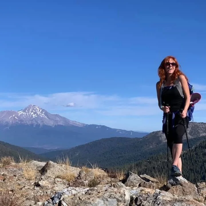 A woman hiking on a rocky trail with a mountain range and snow-capped peaks in the background, under a clear blue sky.