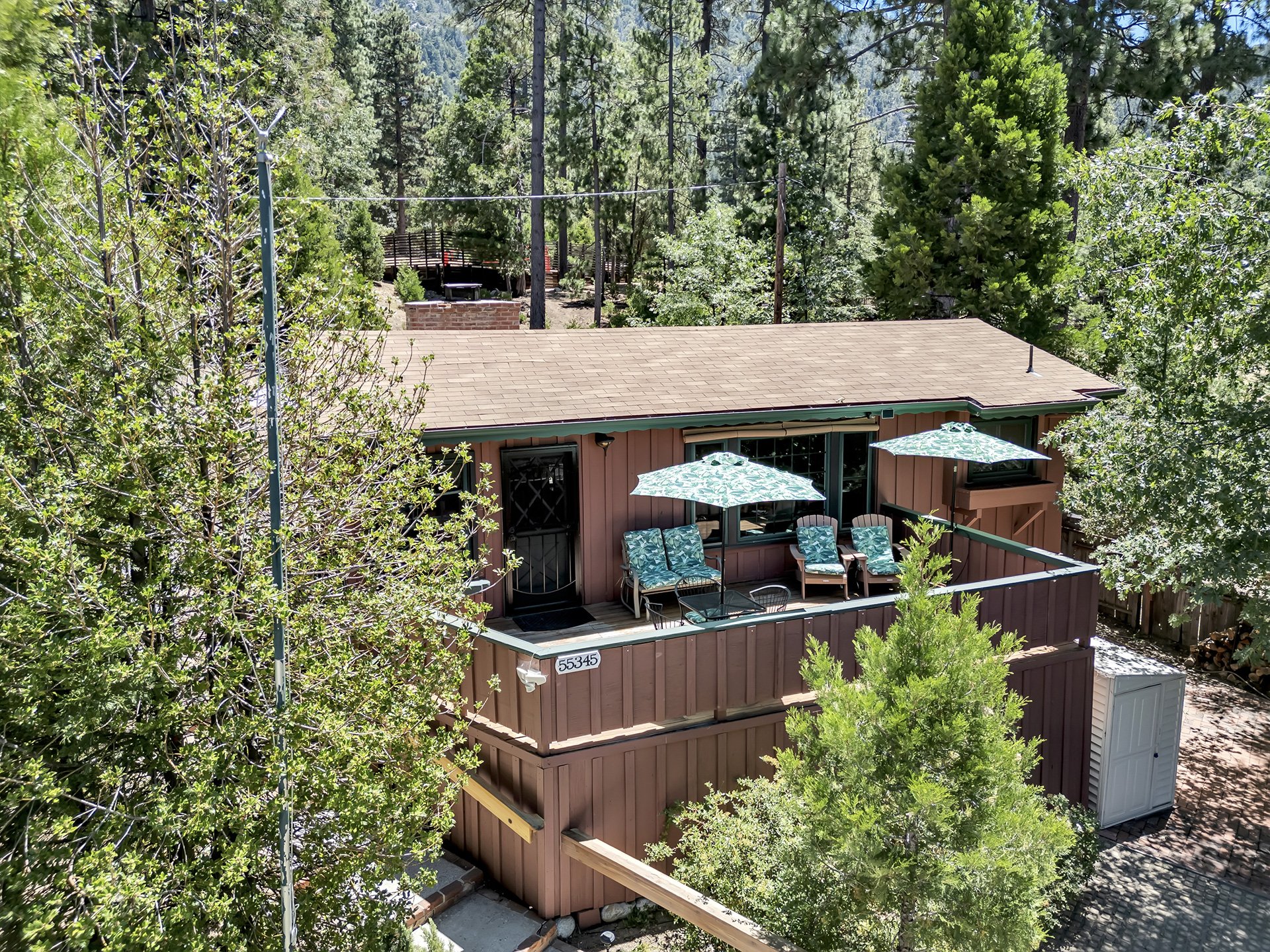 A two-story brown house with a small balcony decorated with patio furniture and umbrellas, surrounded by trees and a forested background.