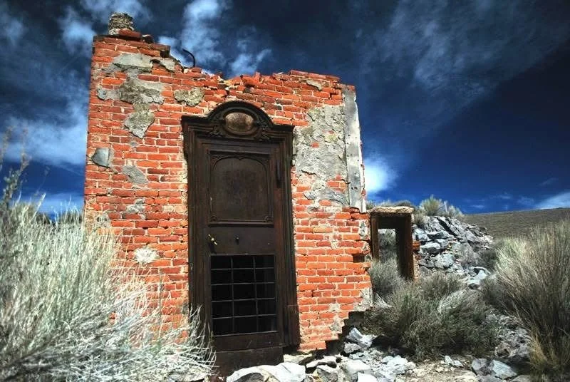 Old brick building with remnants of peeling gray plaster, an arched wooden door with a metal grille at the bottom, and a small window opening on the right. The building is situated in a dry, rocky landscape with sparse grass and shrubs, under a dark,