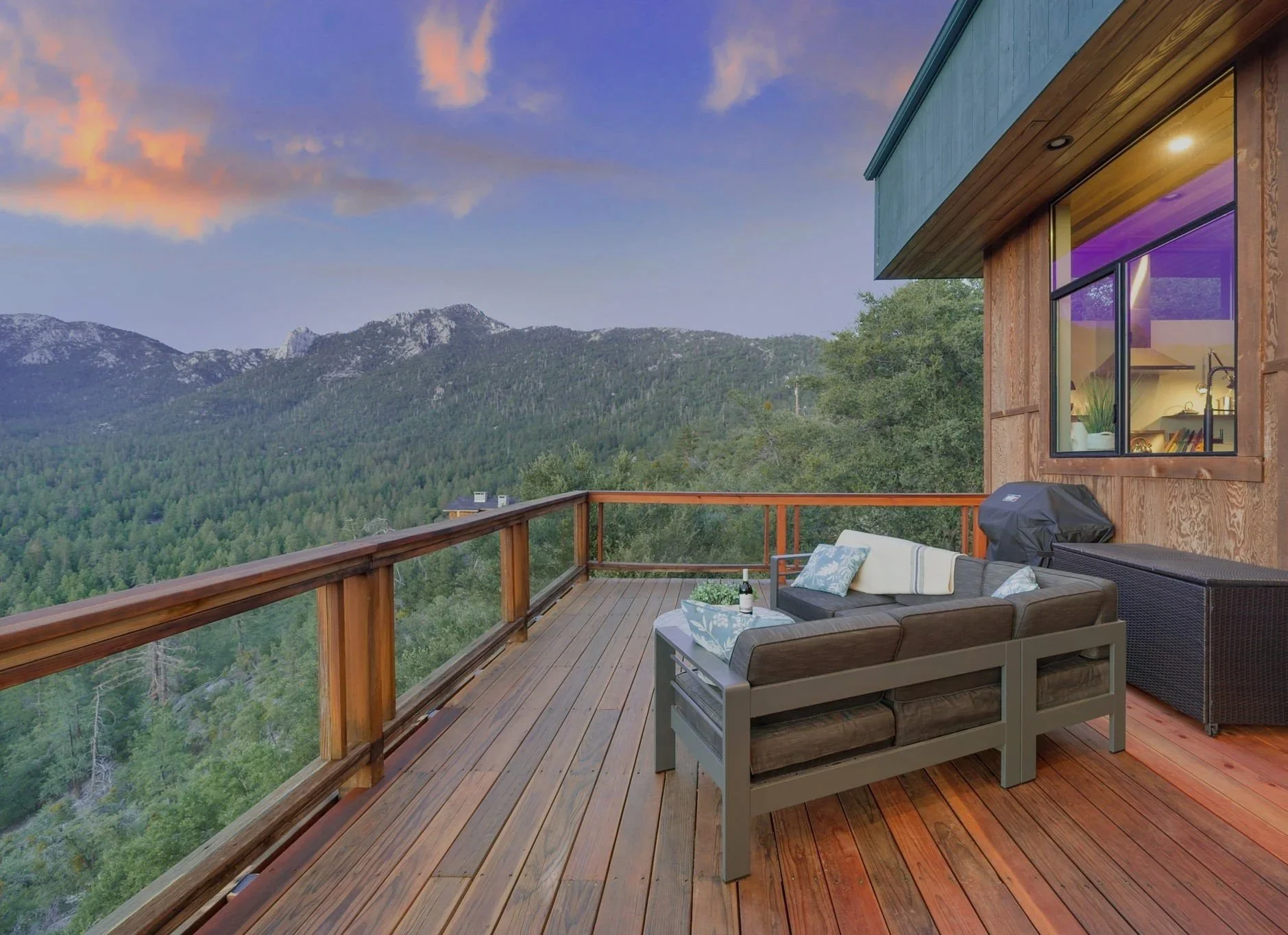 A wooden balcony overlooking a mountain forest at sunset, with a patio sofa, a small coffee table holding a bottle and glasses, and a grill.