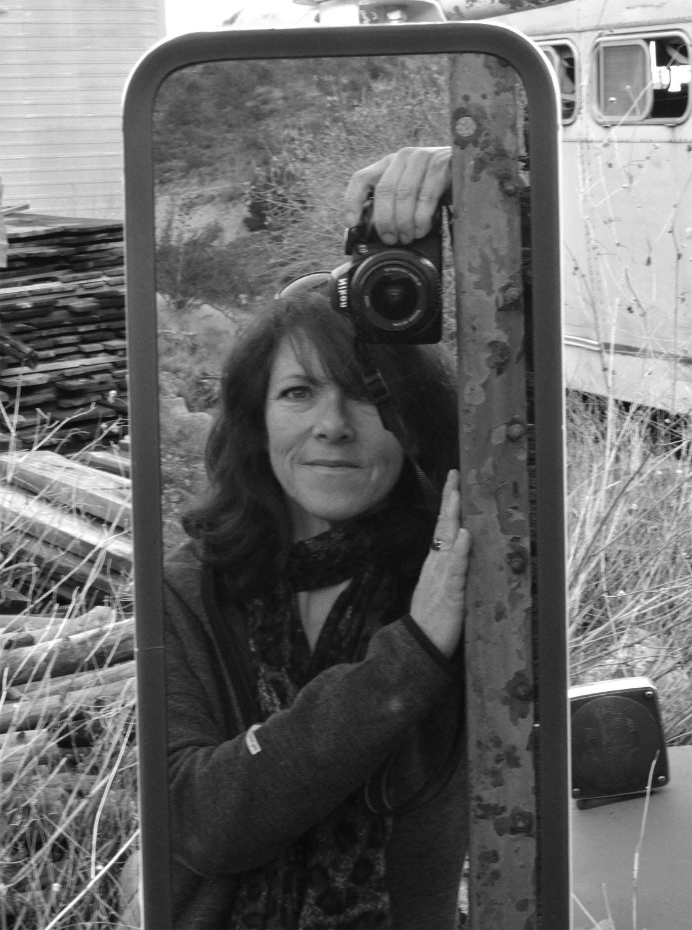 Black and white outdoor scene with a woman taking a selfie using a mirror or reflective surface, holding a Canon camera. The woman has dark hair and is wearing a jacket, with a background of stacked wood and outdoor vegetation.