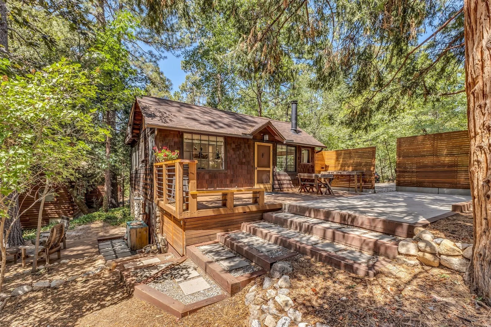 A small wooden house with a deck built into a wooded area with tall trees and a stone pathway leading up to it, surrounded by a wooden privacy fence.