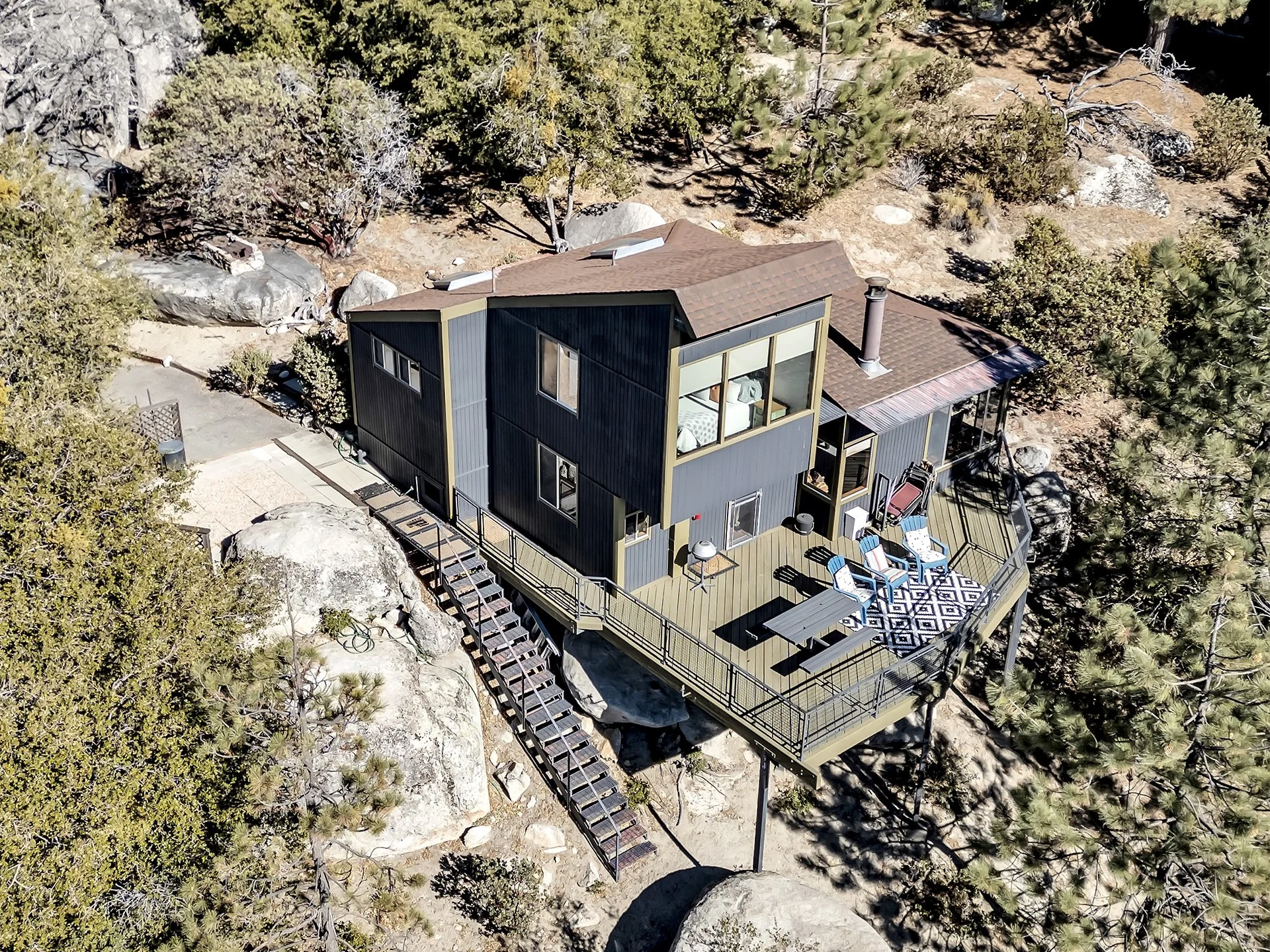 A modern house built on a rocky hillside surrounded by trees, with a large wooden deck and outdoor seating area.
