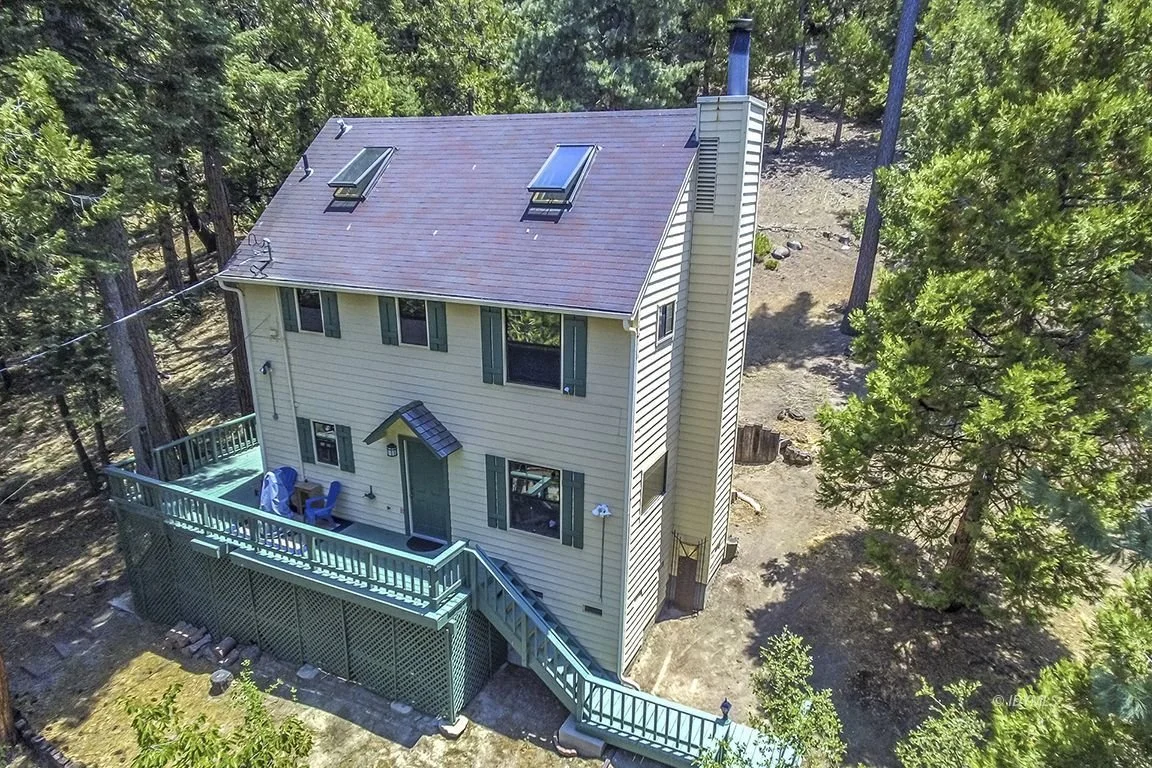 A two-story house with yellow siding, green shutters, and a purple roof, surrounded by trees. The house has a large back deck with outdoor chairs, a staircase, and skylights on the roof.