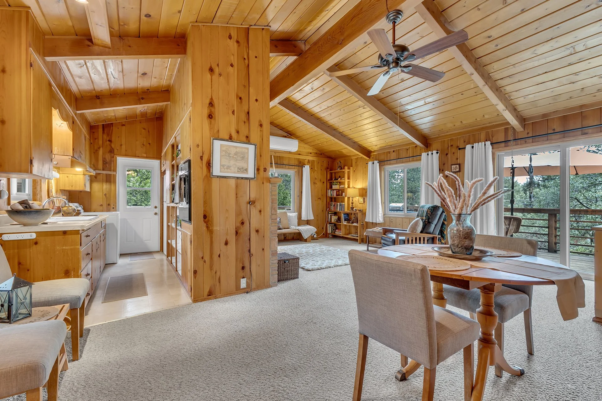 Open-plan living room and kitchen with wood-paneled ceilings, large windows, a ceiling fan, and a dining table with a vase of pampas grass.