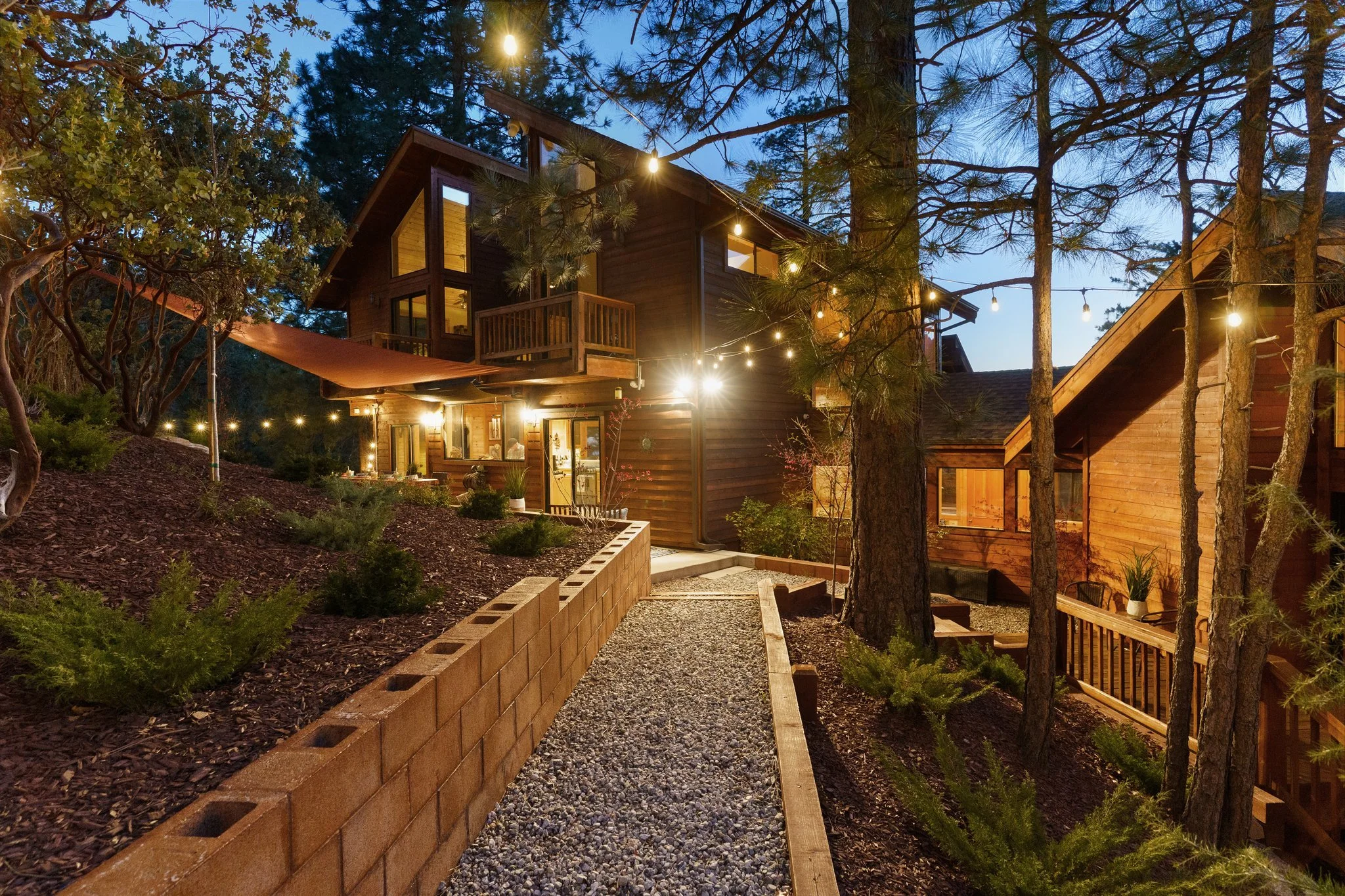 A two-story wooden house surrounded by tall trees at dusk. The house is illuminated with outdoor string lights and interior lights visible through the windows. There is a balcony on the upper floor and a sloped roof. The outdoor area features a gravel pathway, a low brick wall, and landscaped garden beds.