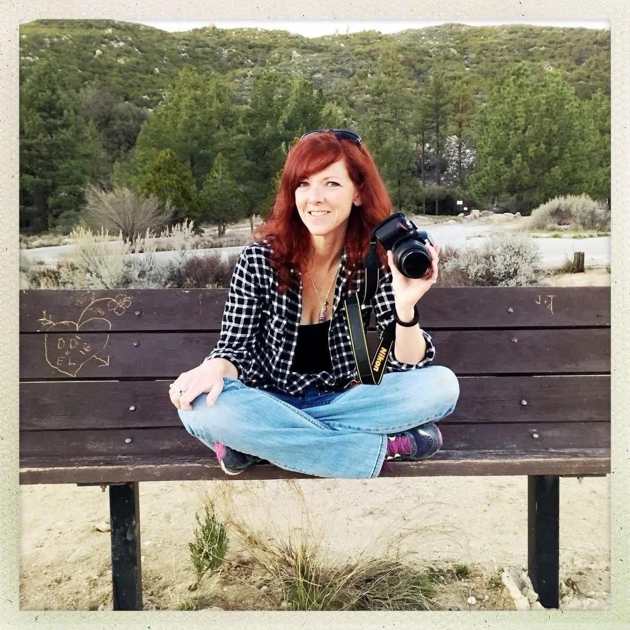 A woman with red hair and sunglasses holding a camera, sitting cross-legged on a wooden park bench in a desert-like area with trees and shrubs in the background.