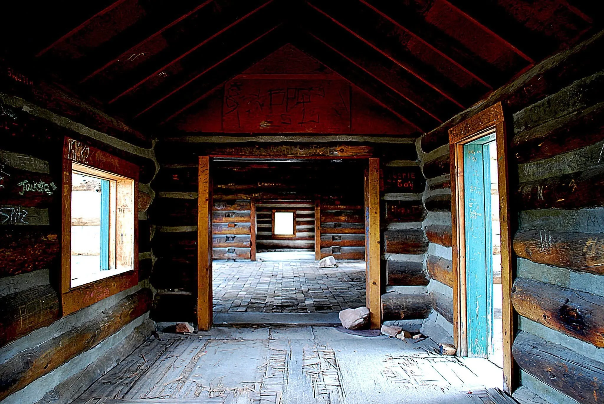 Interior of a rustic log cabin with wooden walls and ceiling, open doorways, and a stone floor, showing a view into another room with log walls and a small window.