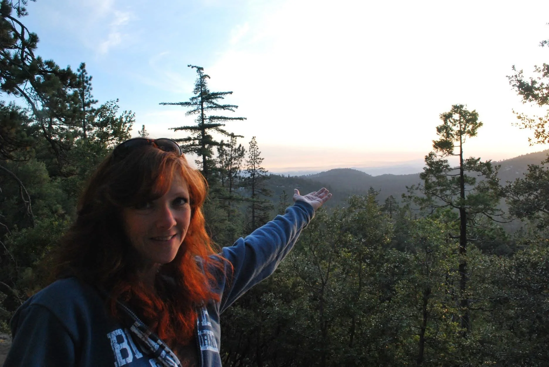A woman with red hair and sunglasses on her head is smiling and pointing at a scenic view of mountains and trees during sunrise or sunset, with a clear sky above.