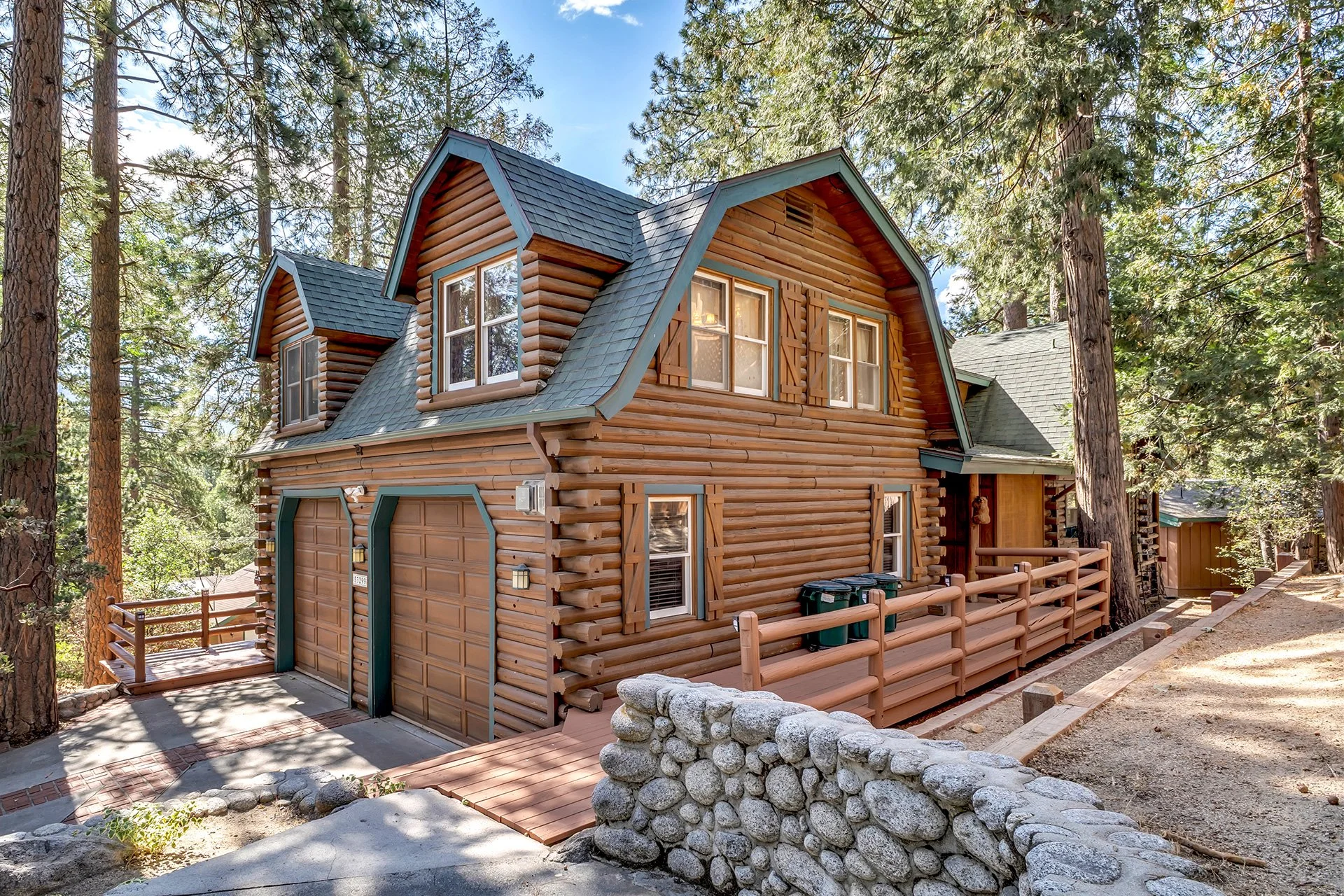 A two-story log cabin house in a forested area surrounded by tall pine trees, with a small deck and a gravel pathway.