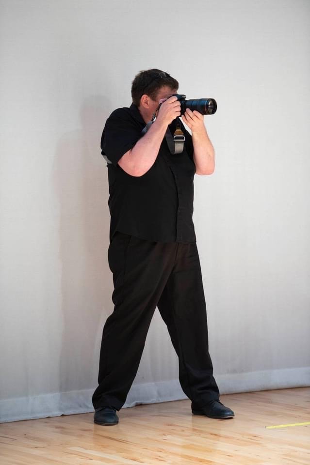 A man dressed in black taking a photograph with a camera against a plain white wall.
