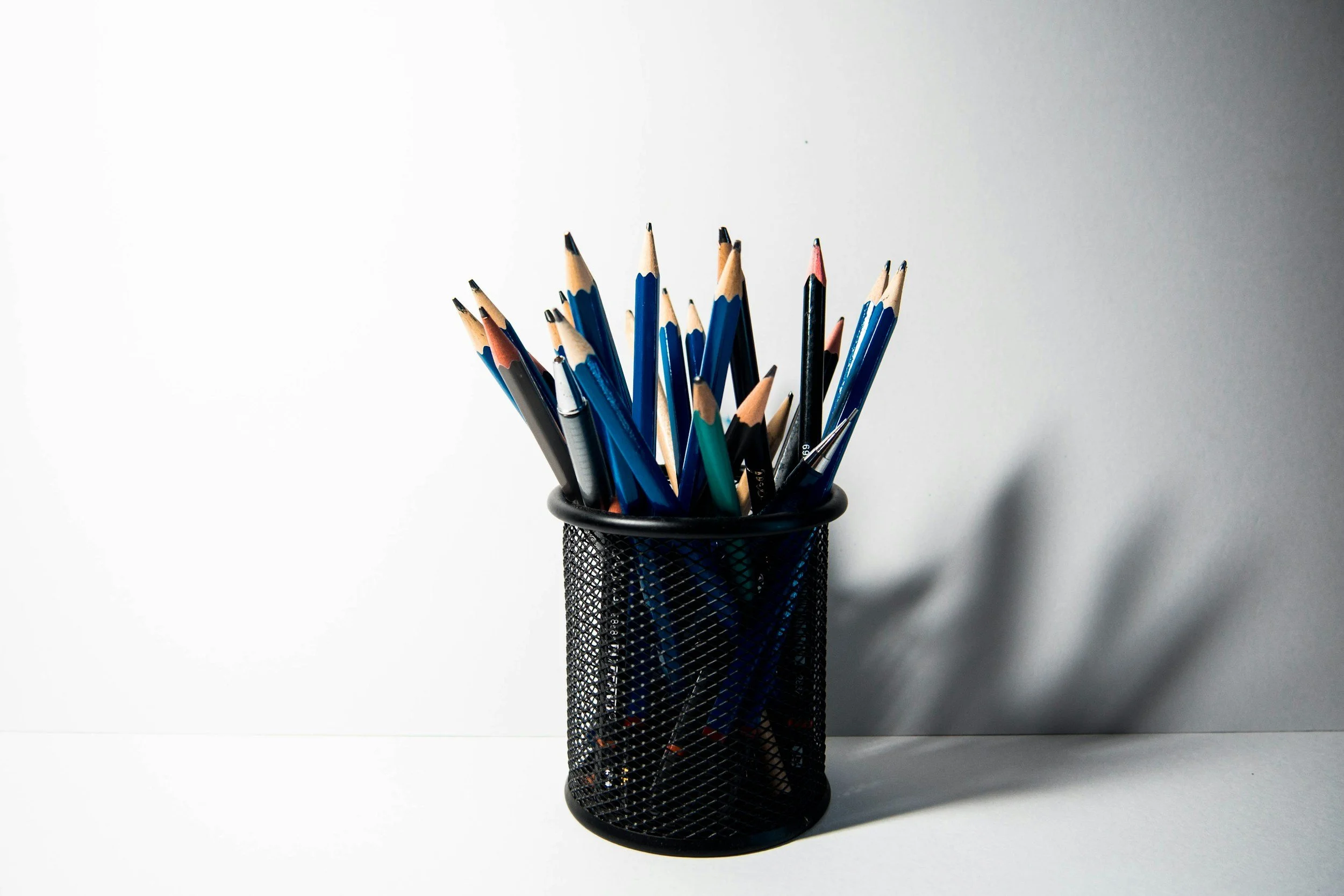 Black metal mesh pen holder filled with blue, black, and colored pencils on a white surface and gray background.