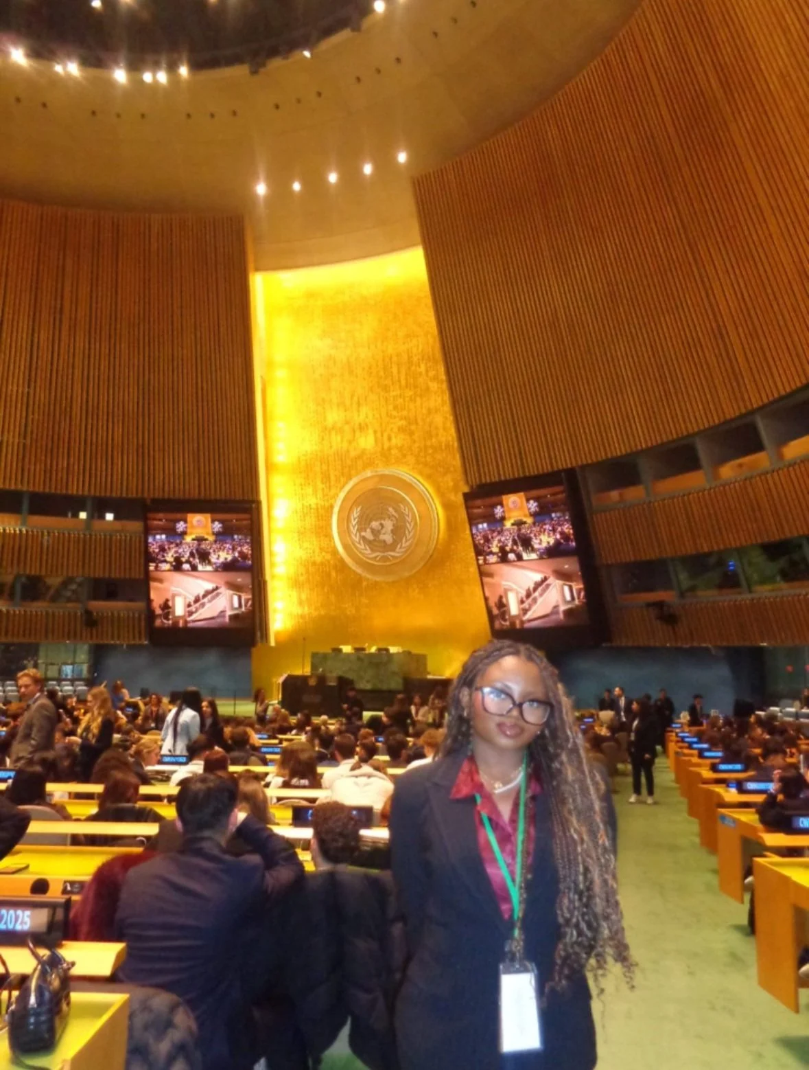 A woman with braids, glasses, and a business suit standing in the United Nations General Assembly hall with many delegates seated at their desks, a large gold emblem and flag on the wall behind her.
