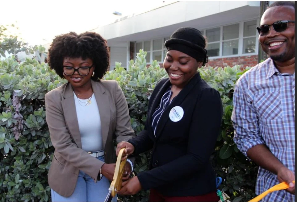 Three people smiling and cutting a ribbon outdoors in front of bushes, celebrating an event.