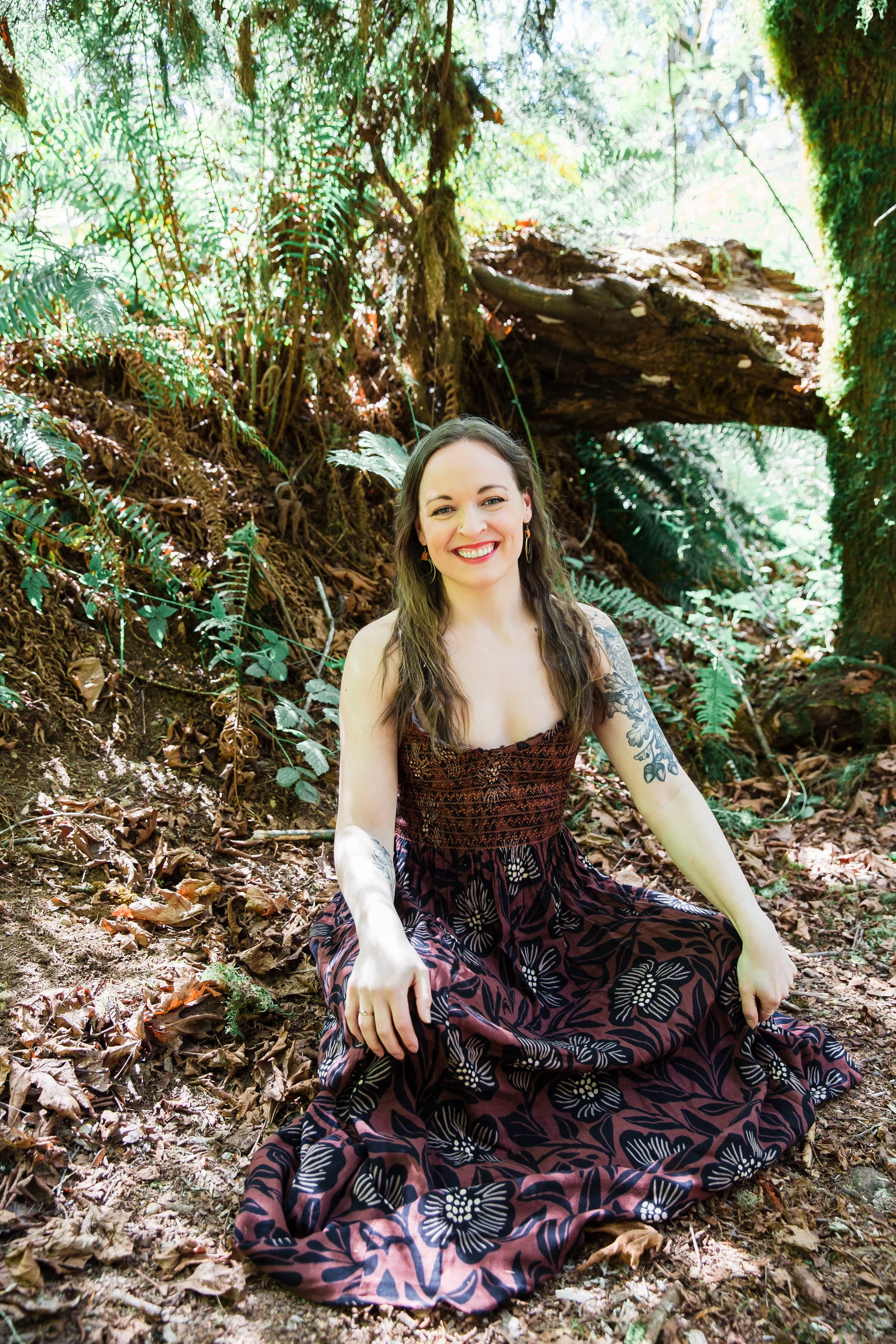 A woman sitting on the forest ground, smiling, surrounded by lush green ferns and trees, wearing a patterned dress.