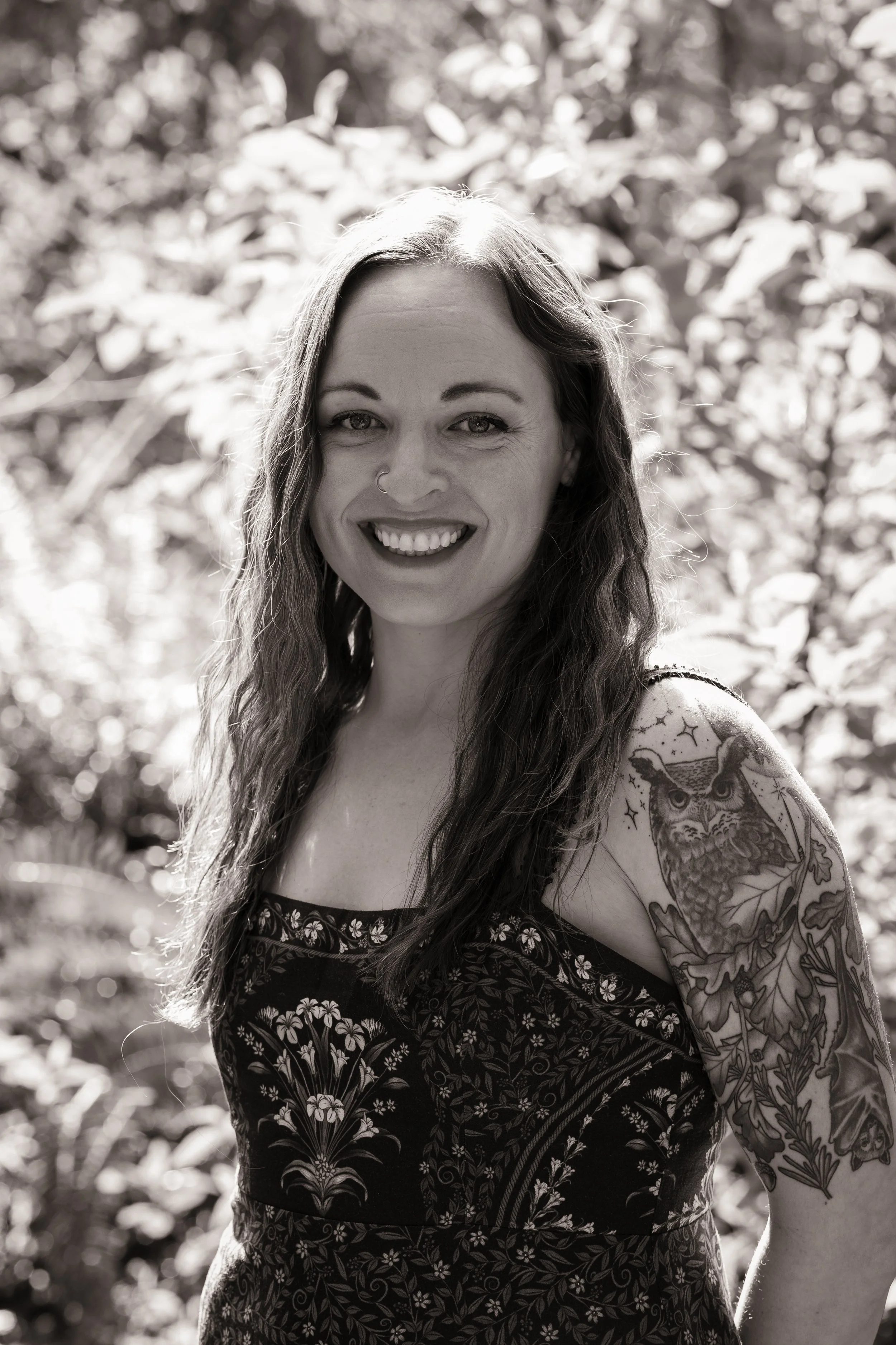 Black and white photo of a smiling woman with long wavy hair, wearing a sleeveless dress with floral patterns, standing outdoors with blurred trees and foliage in the background.