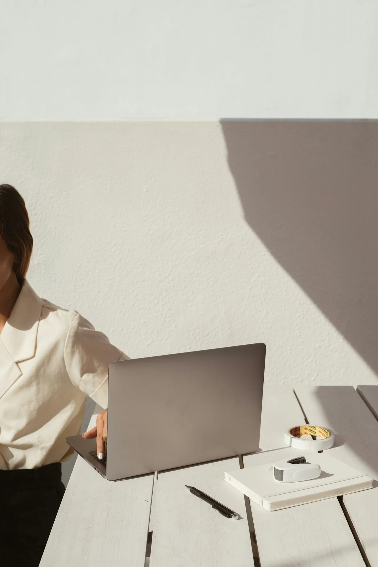 A person sitting at a white desk with a laptop, pen, tape, and a white stapler, with sunlight casting a shadow on the textured wall behind.