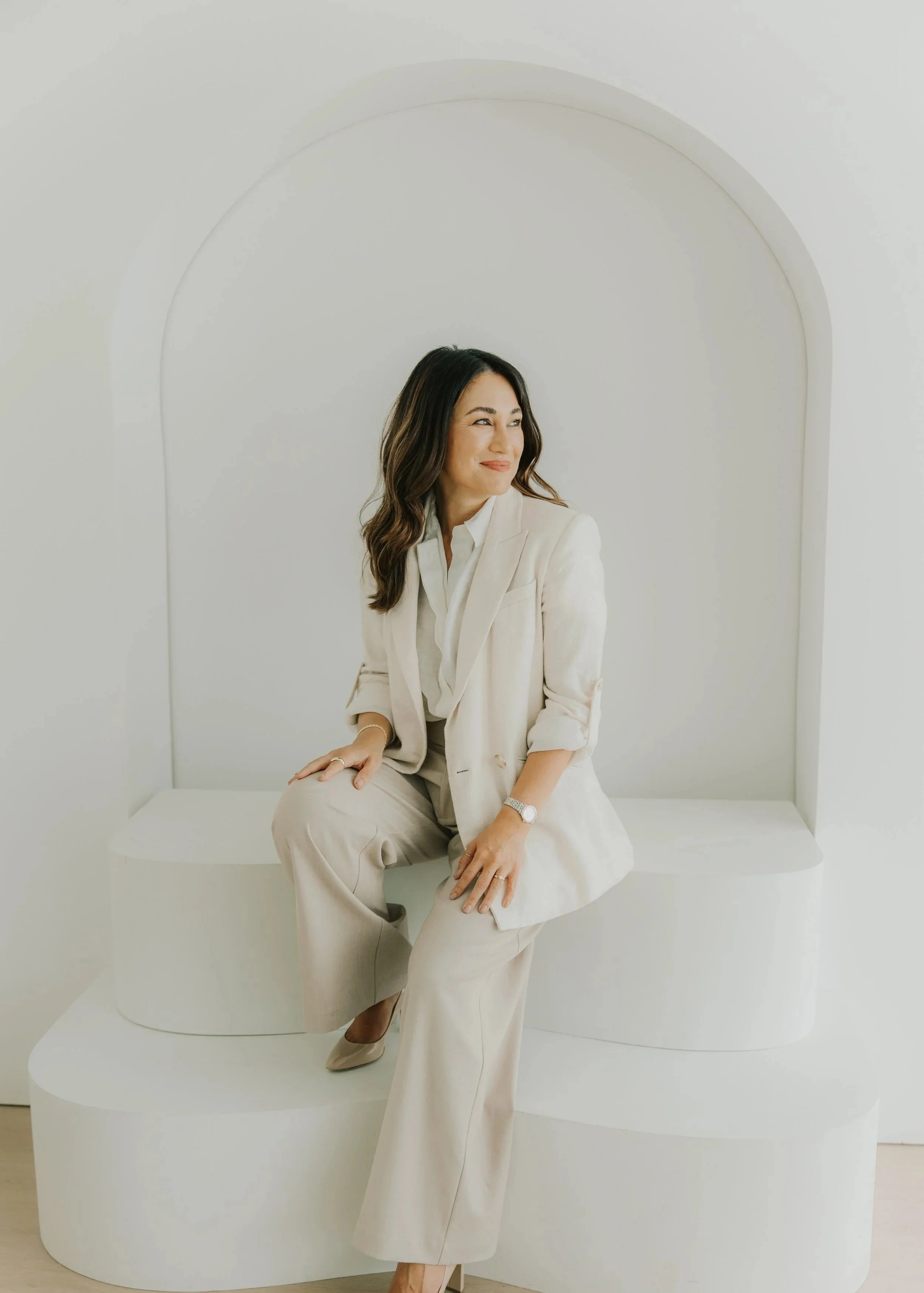 A woman sitting on a white, modern bench with an arched-backrest in a minimalistic white room. She is wearing a beige suit and smiling with a playful expression.