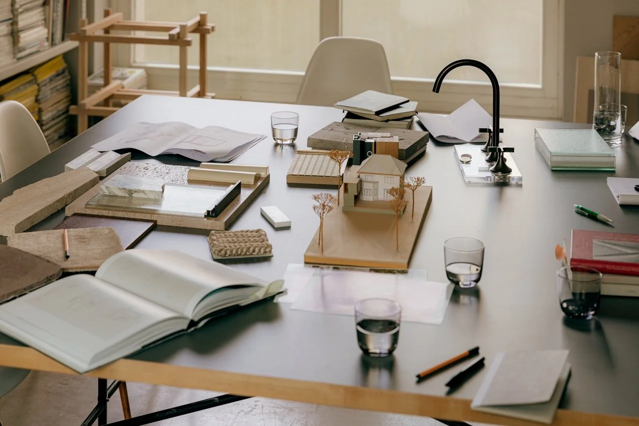 Design studio workspace with architectural models, books, papers, markers, and glasses of water on a table, with shelves and a window in the background.