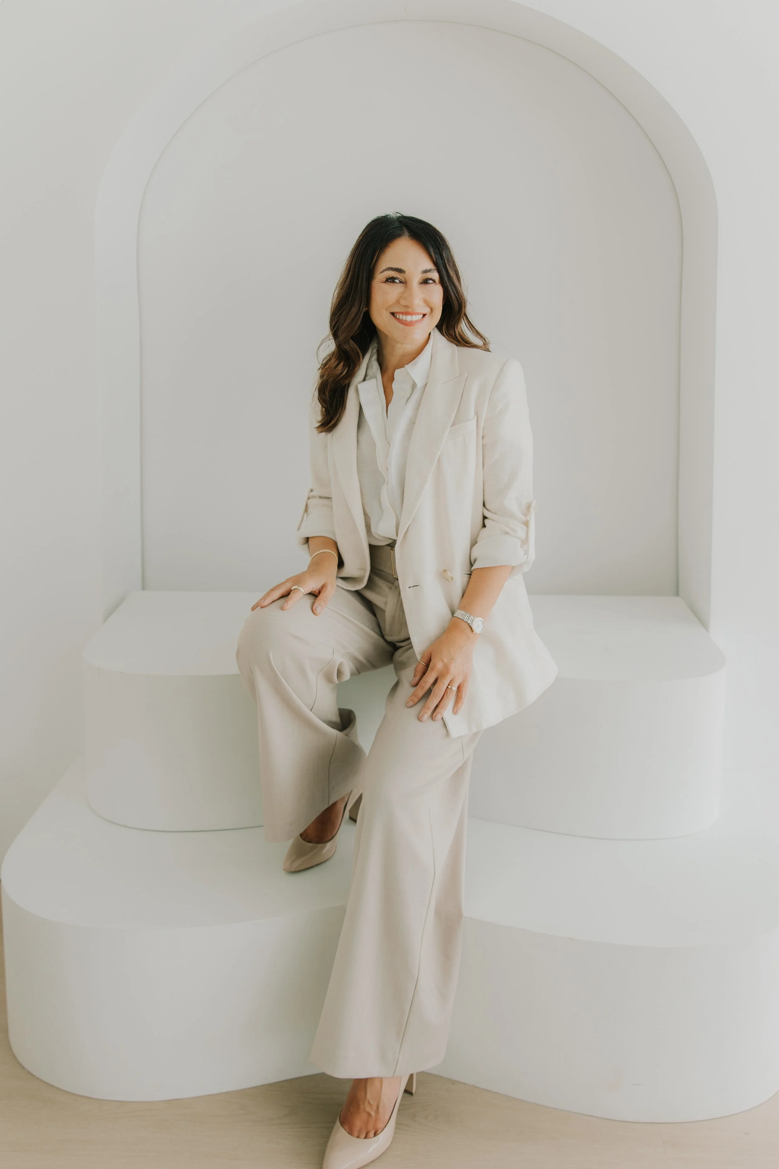 Woman in a cream suit sitting on a white platform against a white background, smiling at the camera.