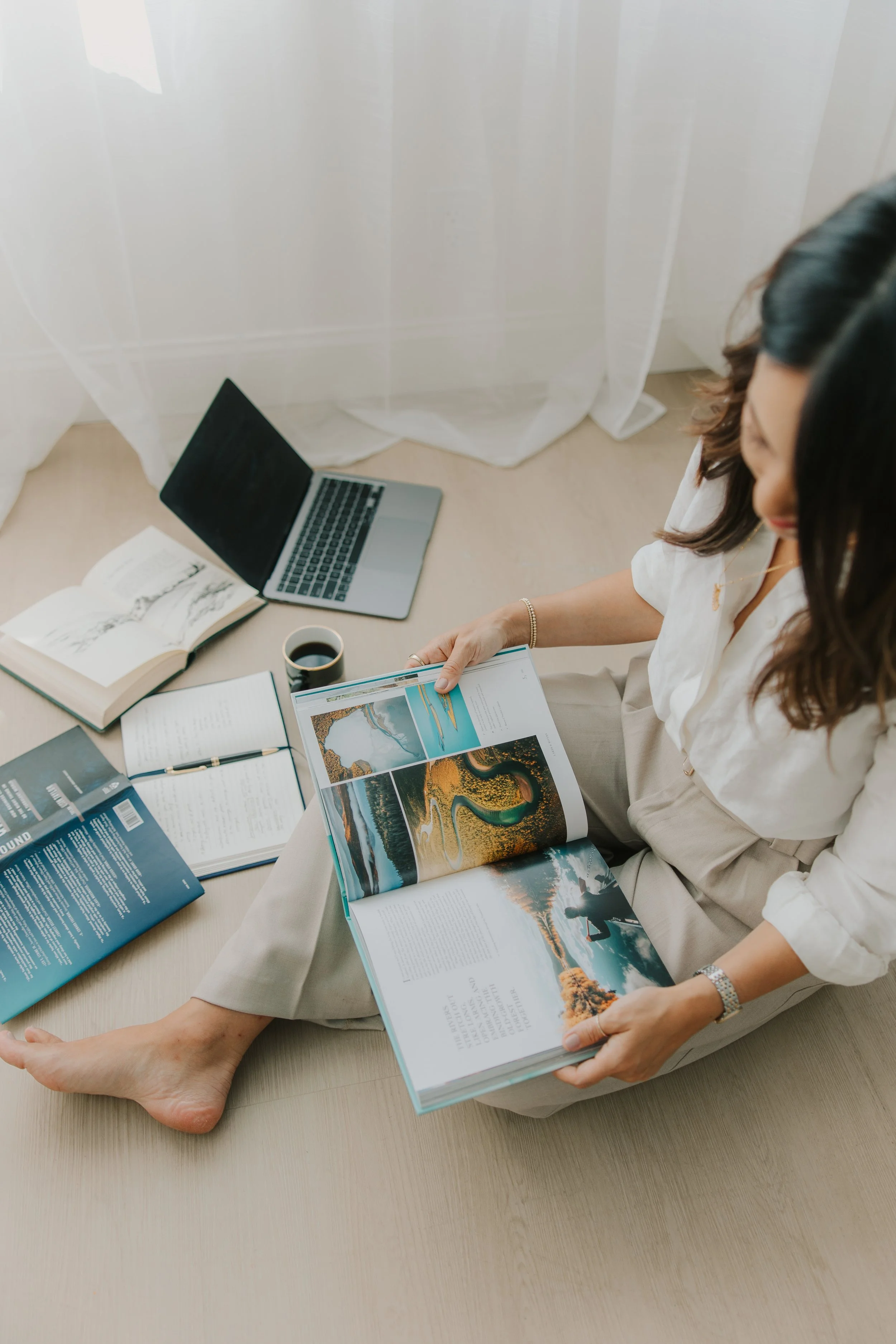 A woman sitting on the floor reading a large coffee table book with colorful landscape photos, surrounded by a laptop, cups of coffee, notebooks, and a pen.