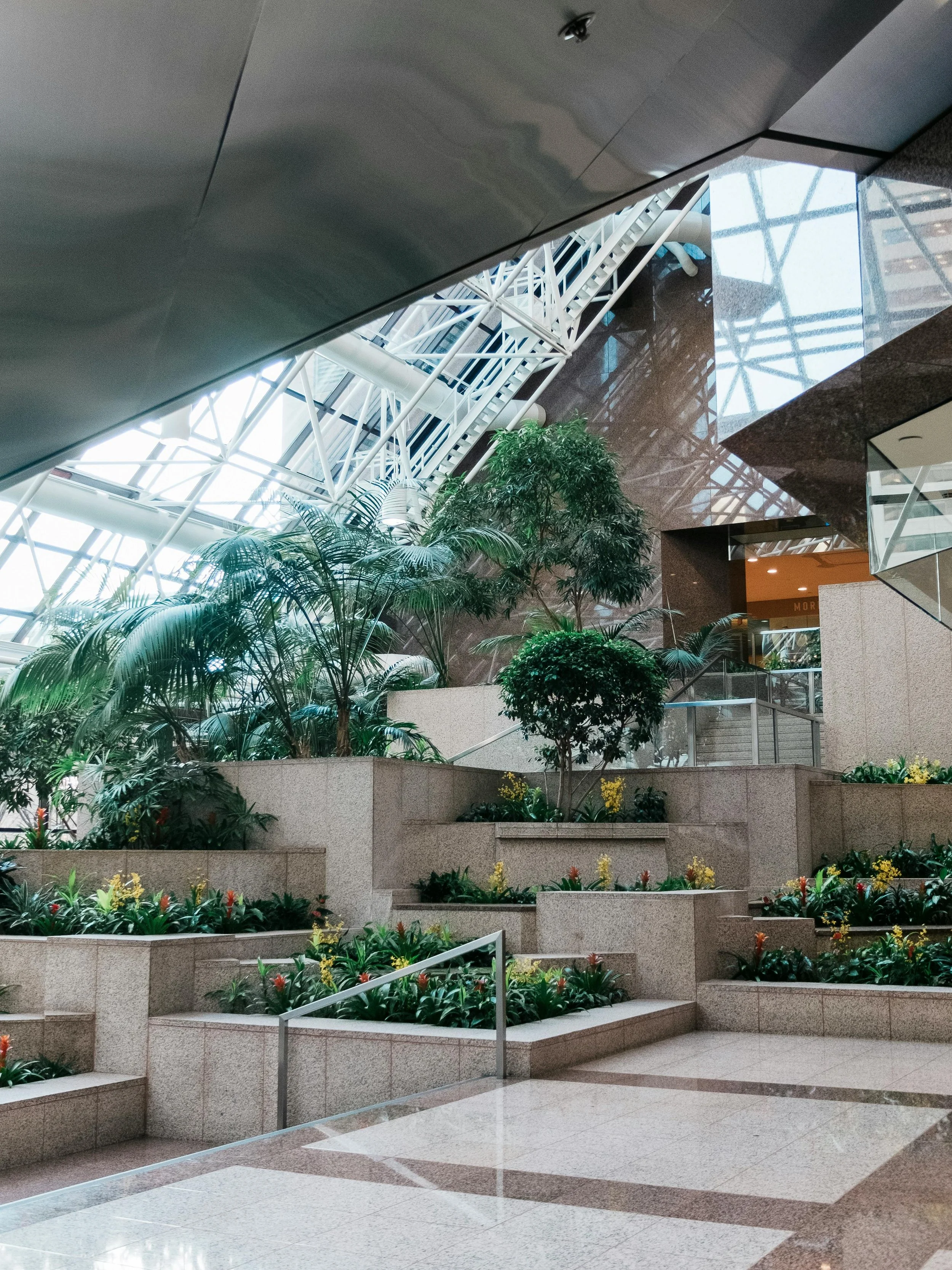 Interior view of a modern shopping mall with escalators, plants, and stairs leading up to different levels.